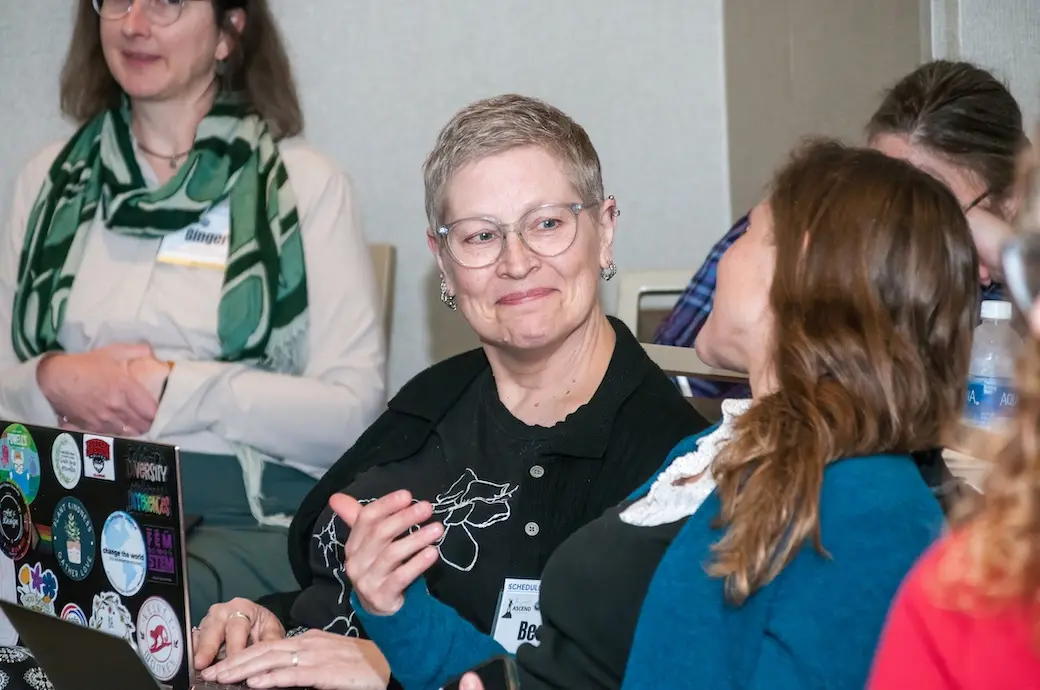 Woman sitting indoors looking at another woman who is turned toward her. Women in background listening.