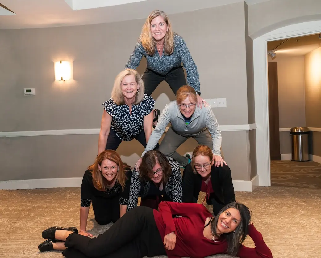 Seven women forming a human pyramid indoors; 3 on bottom row, 2 in middle, 1 on top, with 1 lying on the floor in front.