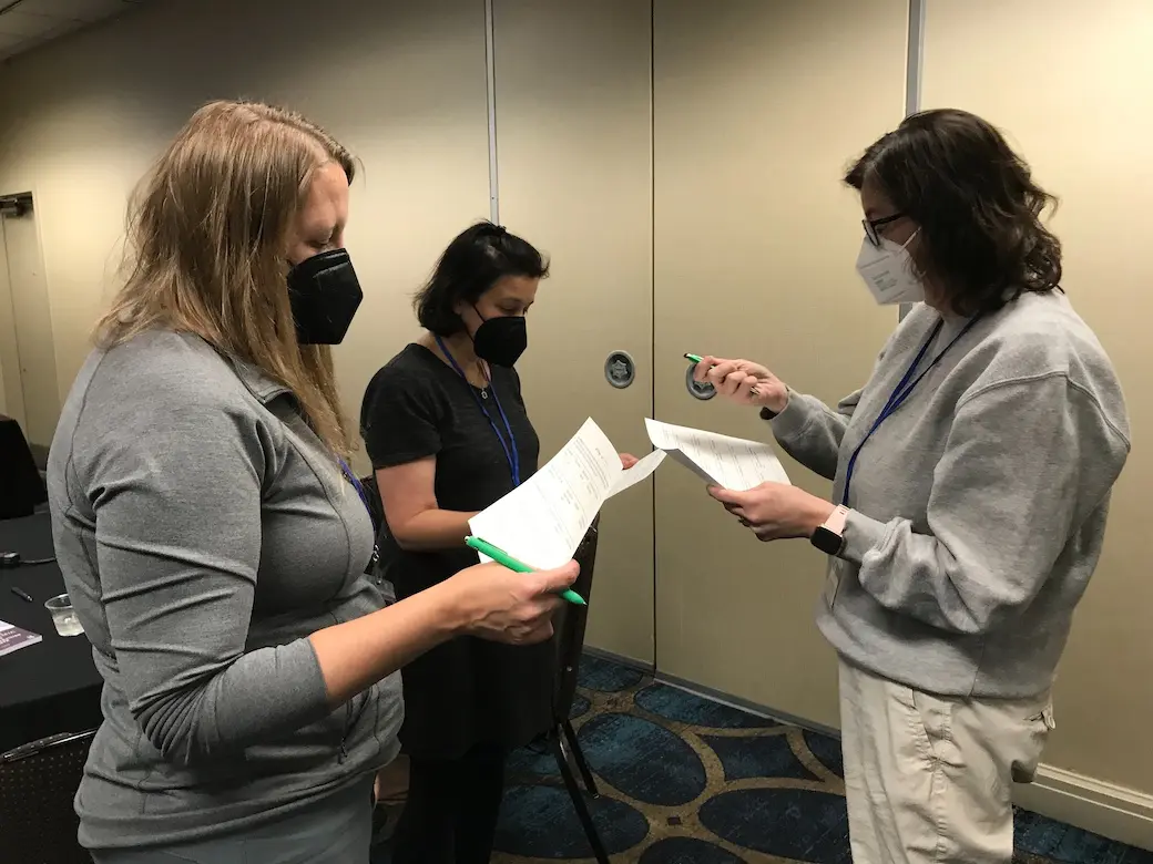 Three women indoors wearing masks standing facing each other reading from papers in their hands.