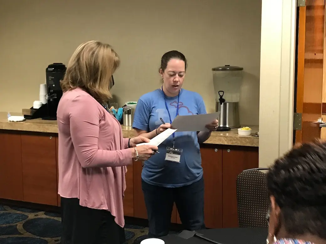 Two women indoors standing turned toward each other reading from papers in their hands.