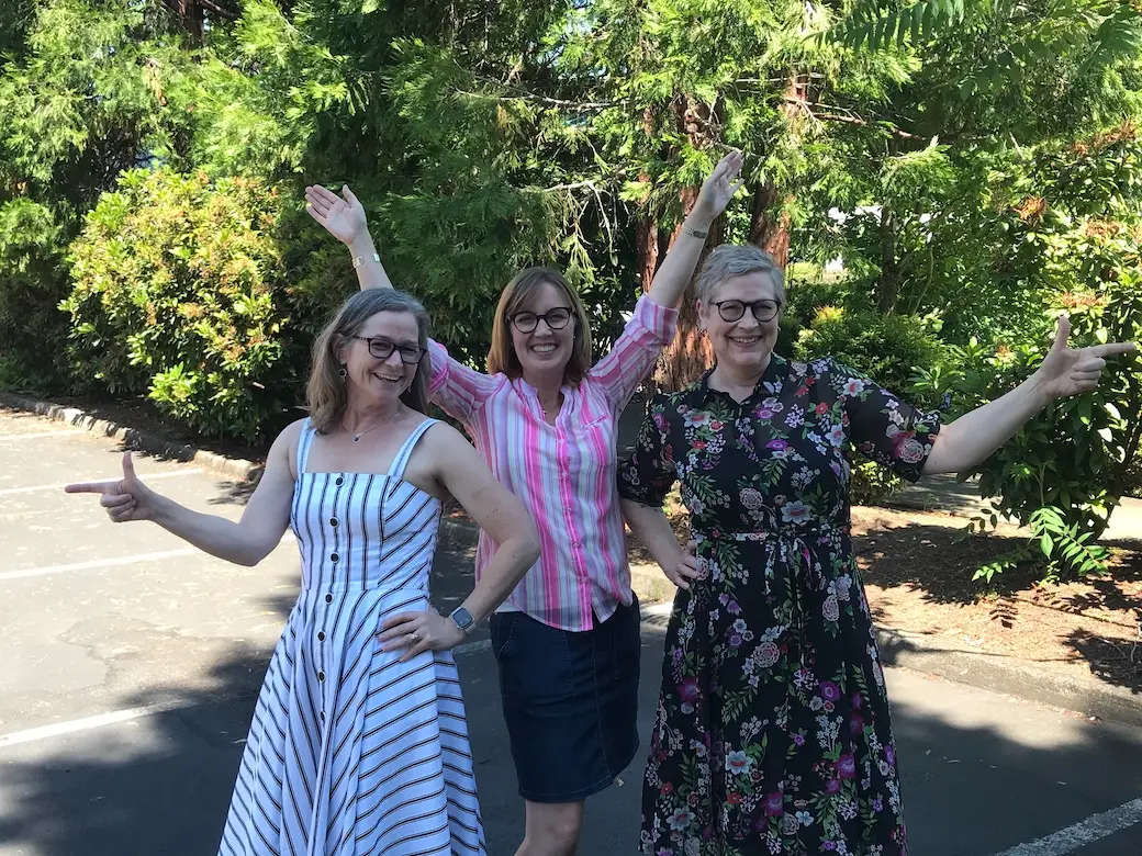 Three women standing outside in front of trees smiling in “Charlie's Angels” pose.