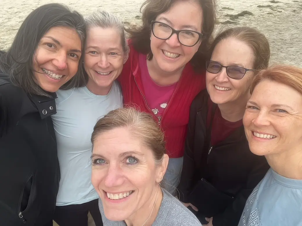 Selfie of five smiling women standing, forming a semicircle around a smiling woman on a beach with sand in background.