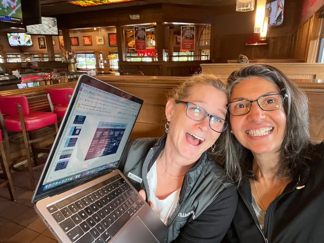 Two women smiling with heads together sitting in a restaurant booth; one holds up laptop computer to show part of the screen.