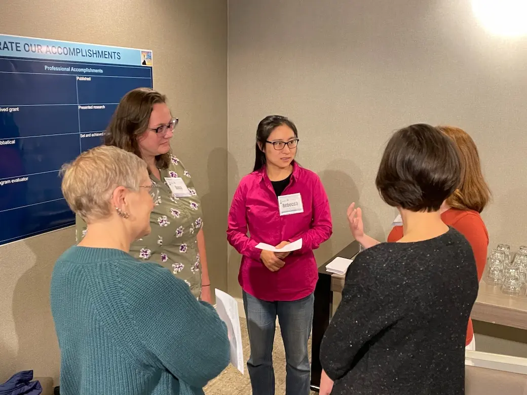 Five women standing in a circle looking at one of them speaking and holding papers in a corner of a hotel conference room.