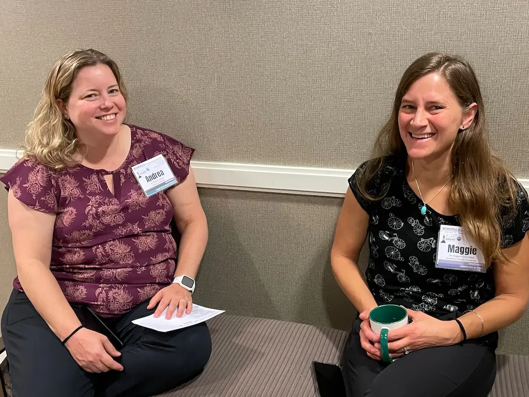 Two women smiling and sitting on a bench with a space between them in a hotel conference room.