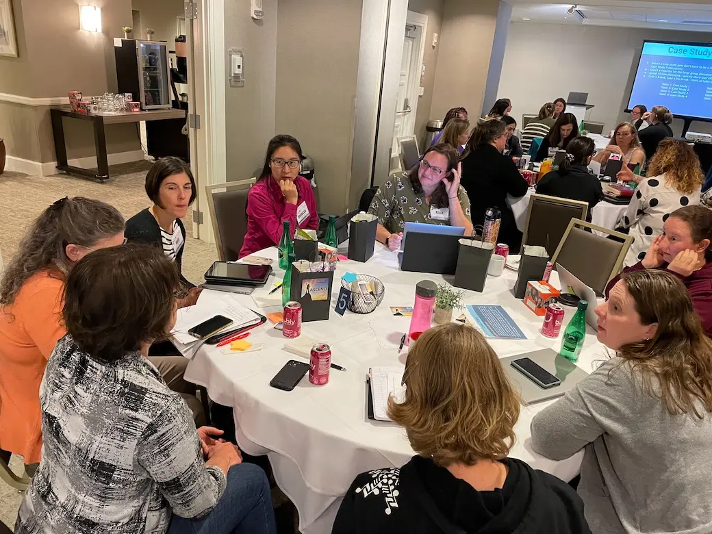 Birds eye view of seven women sitting at table talking using computers and another table of women in background.