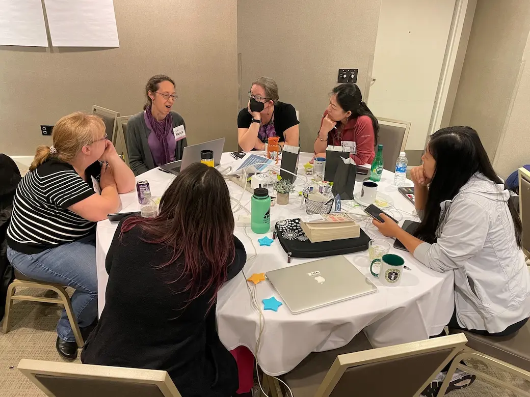 Six women sitting around a table with computers listening to one speak.