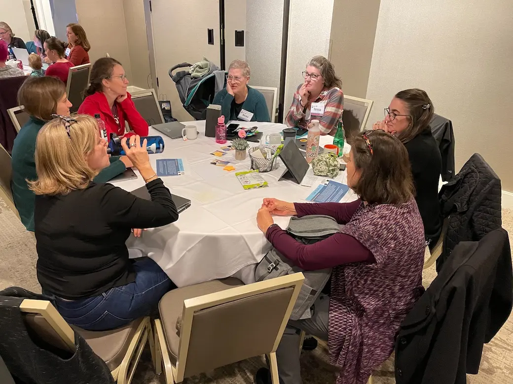 Seven women sitting around a table in a hotel conference talking.