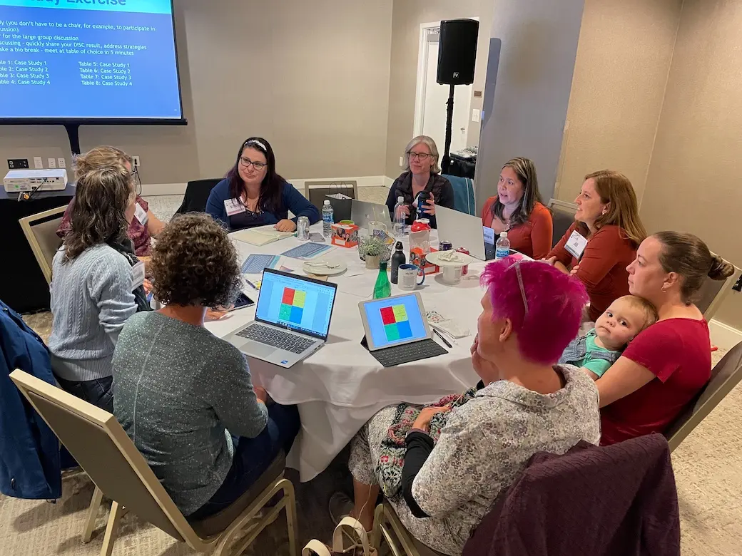 Nine women sitting around a table with computers open talking in a hotel conference room. One is holding a baby. 