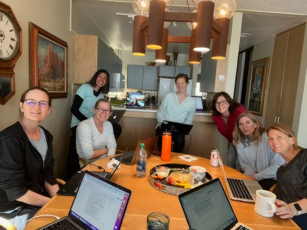 Seven women sitting around a dining room table with open computers taking a break from working.