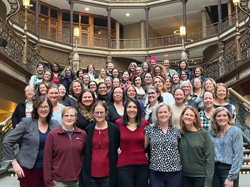 A group of approximately 60 women standing on a grand staircase in an ornate building smiling at the camera.