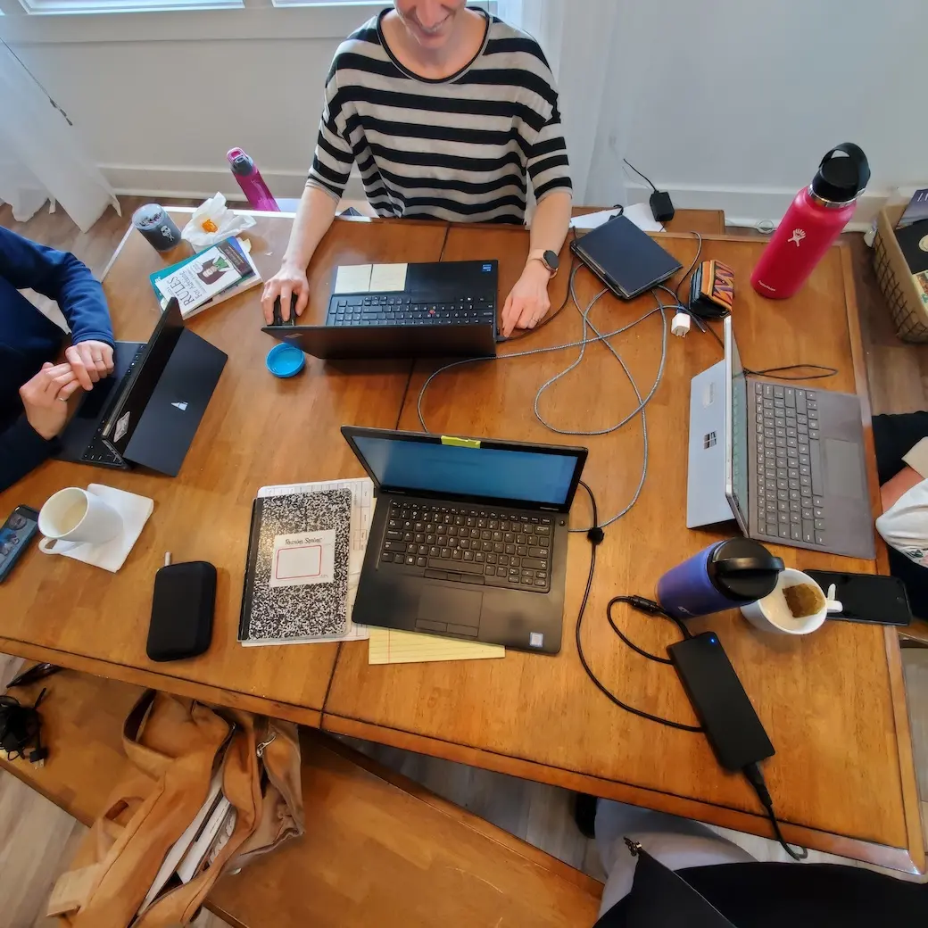 Birds eye view of a table with four laptops, notebooks and cups with one woman's torso in view.