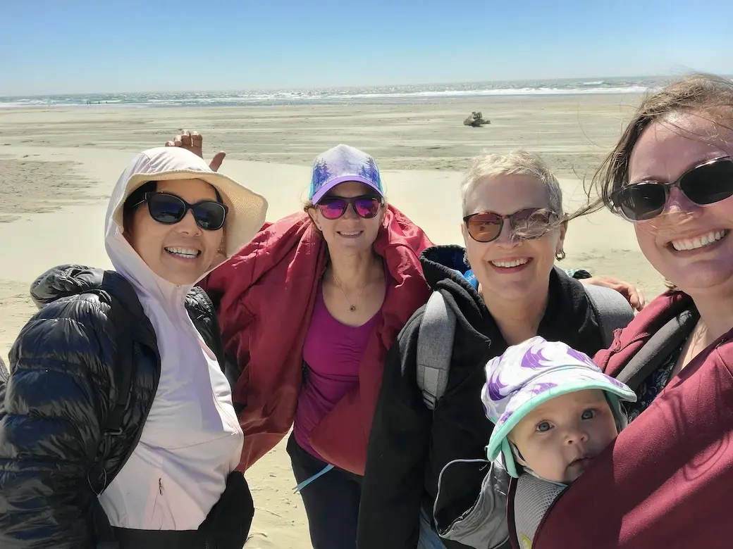 Four women, with one holding a baby in the front carrier, wearing sunglasses and coats standing on a beach.